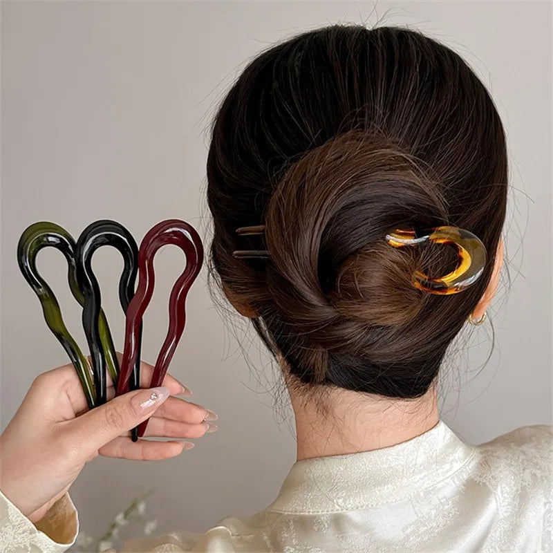 Low twisted bun held by a tortoiseshell-pattern French hair pin, with olive, black and burgundy pins in hand
